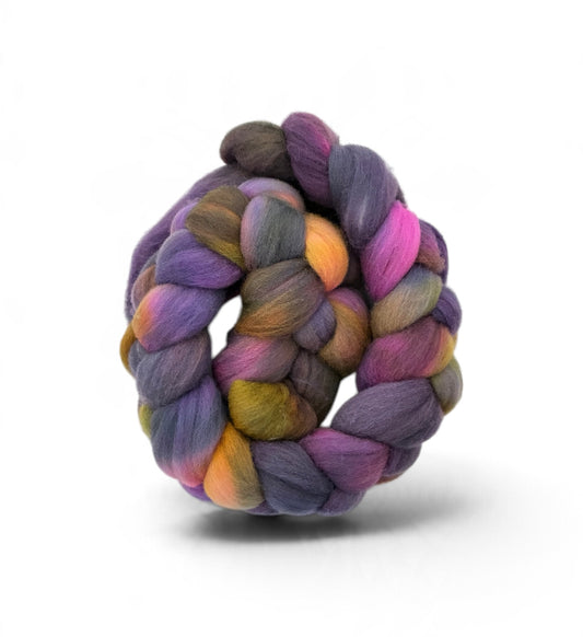 A coiled skein of variegated wool fiber in blended purple, pink, orange, yellow, and gray tones, photographed on a clean white background with a soft shadow.