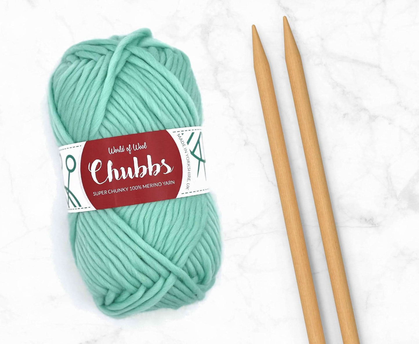Turquoise yarn ball of super chunky 100% merino wool with World of Wool 'Chubbs' label and wooden knitting needles on a white background
