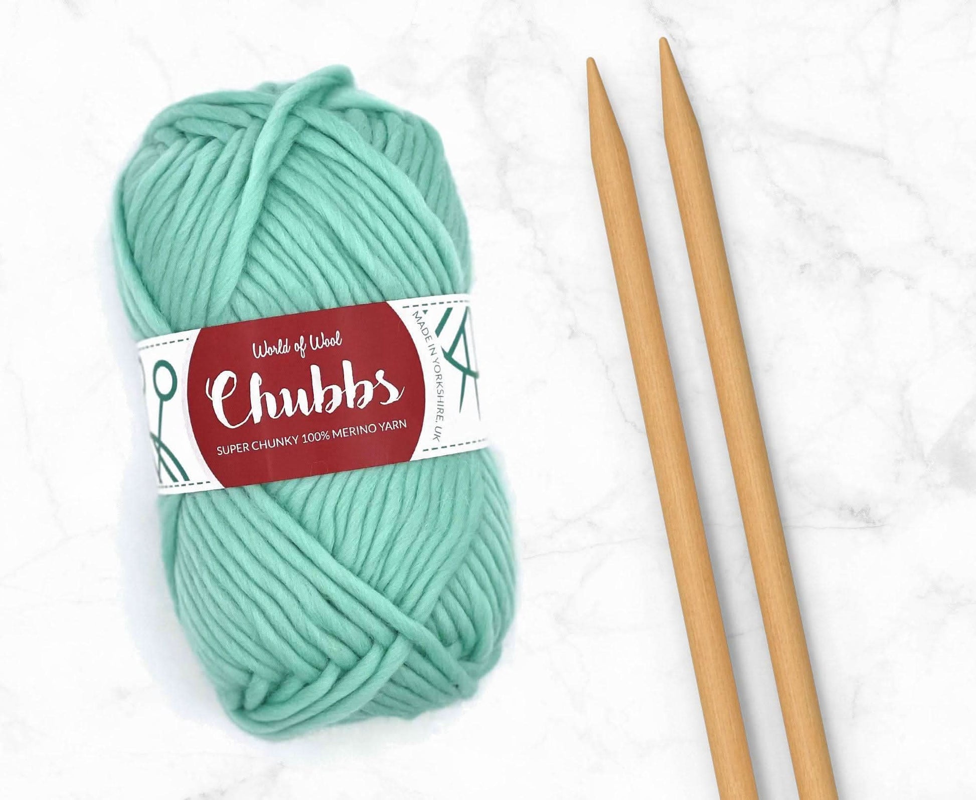 Turquoise yarn ball of super chunky 100% merino wool with World of Wool 'Chubbs' label and wooden knitting needles on a white background
