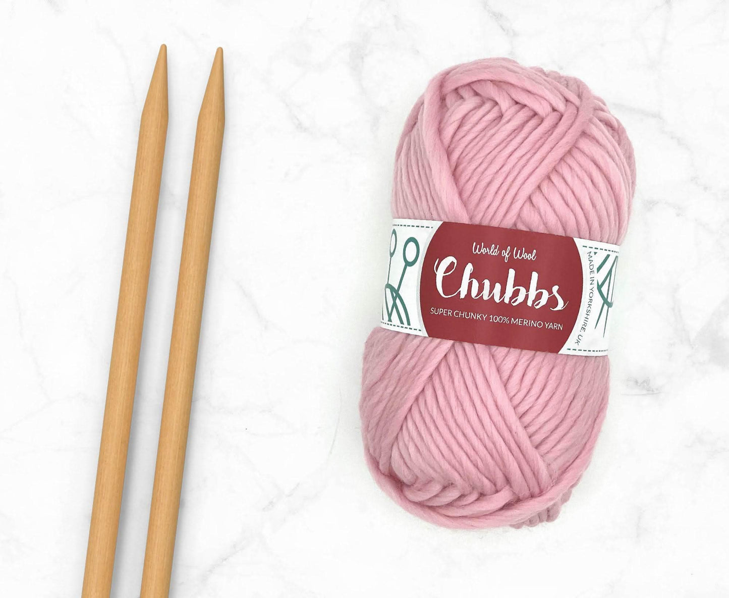 Pink yarn ball of super chunky 100% merino wool with World of Wool 'Chubbs' label and wooden knitting needles on a white background