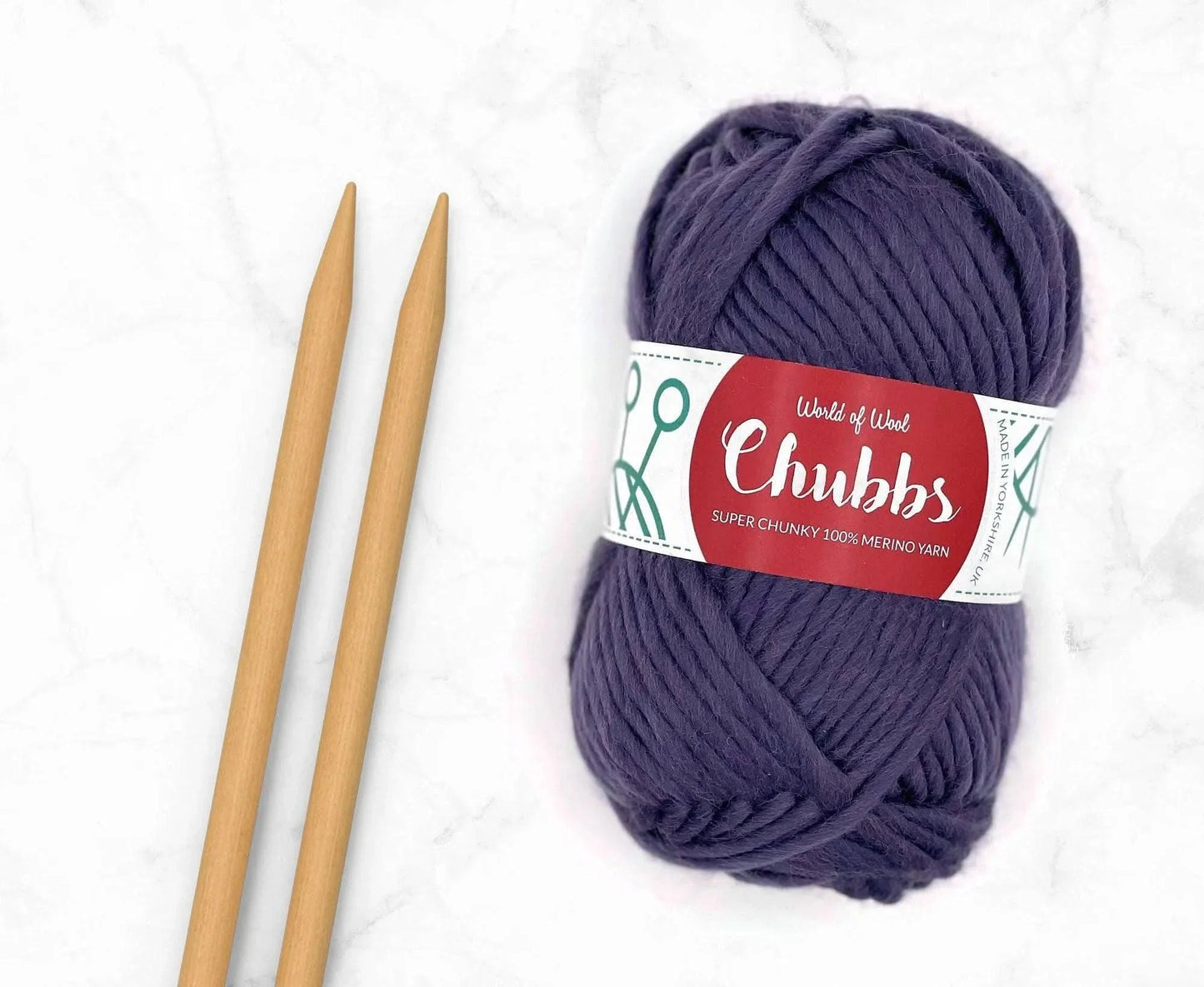 Purple yarn ball of super chunky 100% merino wool with World of Wool 'Chubbs' label and wooden knitting needles on a white background