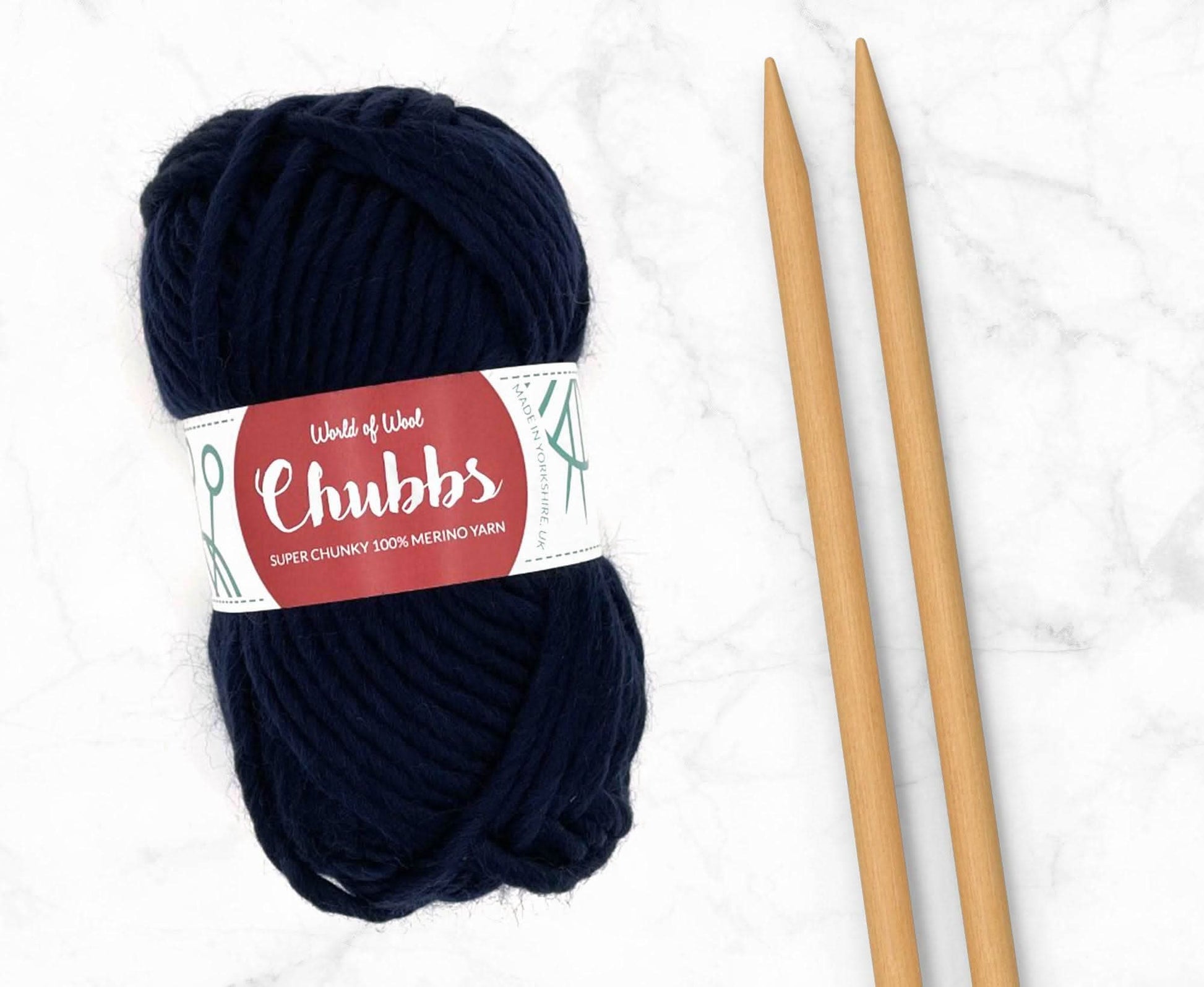 Navy yarn ball of super chunky 100% merino wool with World of Wool 'Chubbs' label and wooden knitting needles on a white background