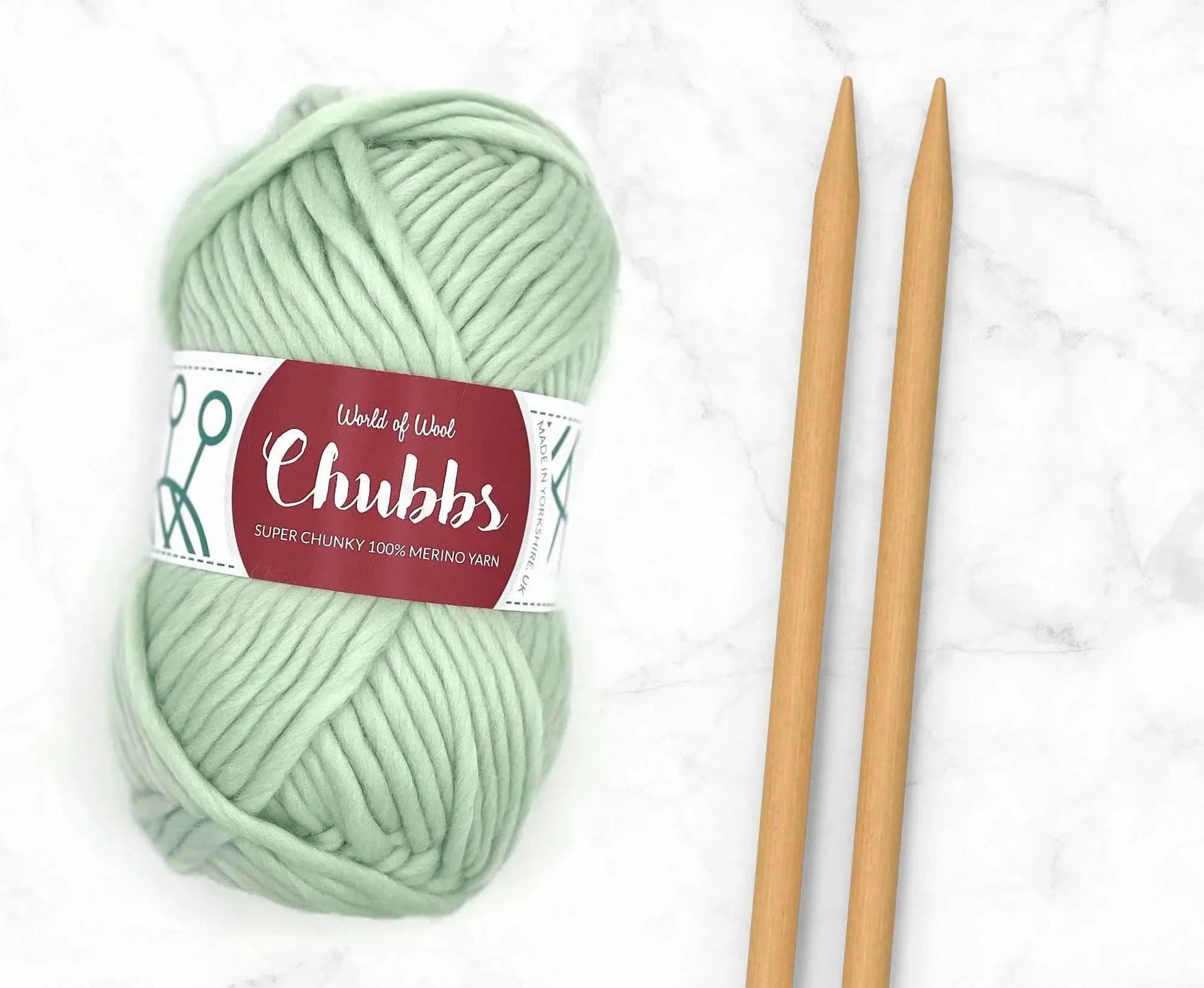 Light green yarn ball of super chunky 100% merino wool with World of Wool 'Chubbs' label and wooden knitting needles on a white background