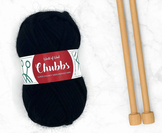 Black yarn ball of super chunky 100% merino wool with World of Wool 'Chubbs' label and wooden knitting needles on a white background