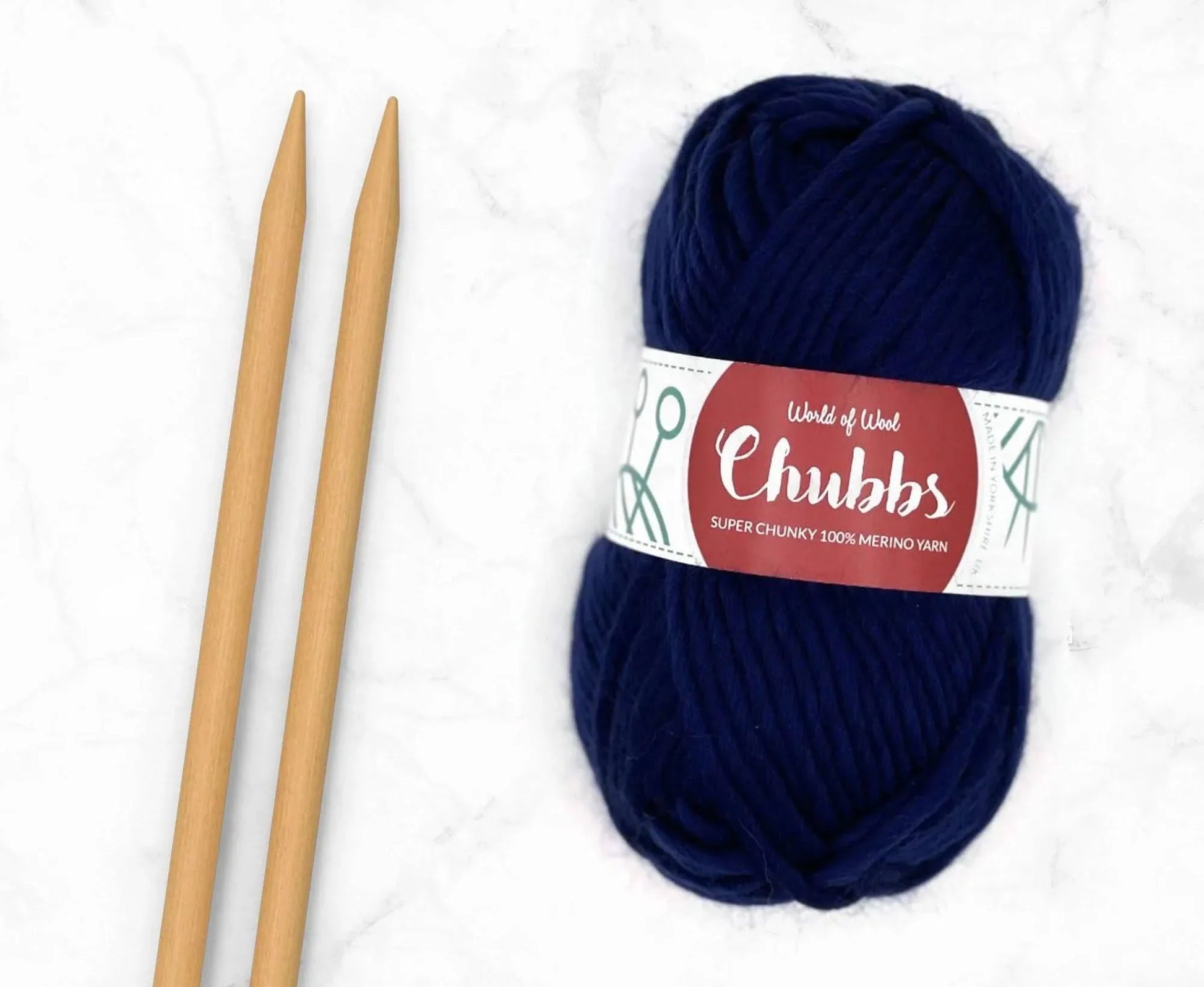Navy yarn ball of super chunky 100% merino wool with World of Wool 'Chubbs' label and wooden knitting needles on a white background