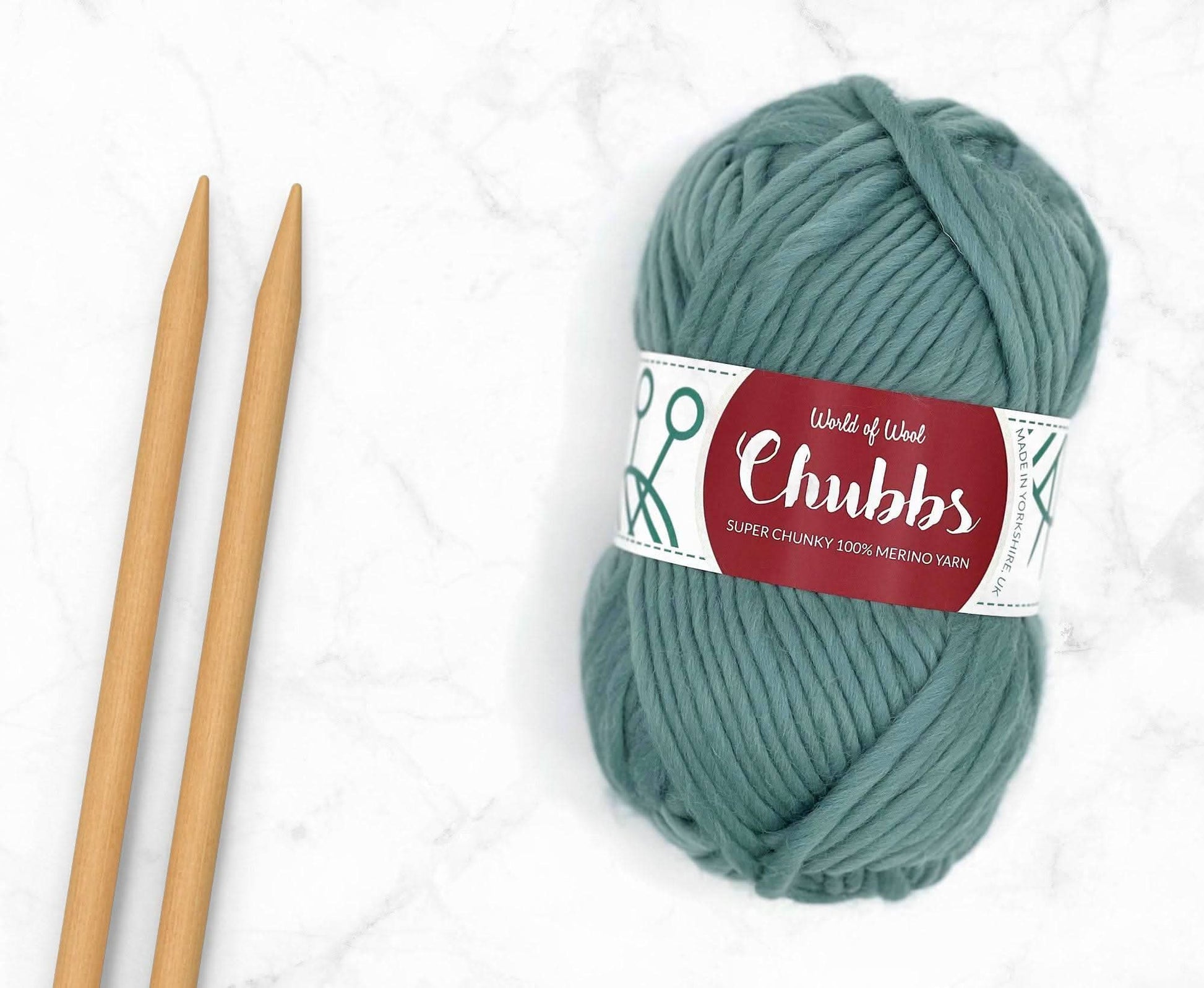 Teal yarn ball of super chunky 100% merino wool with World of Wool 'Chubbs' label and wooden knitting needles on a white background